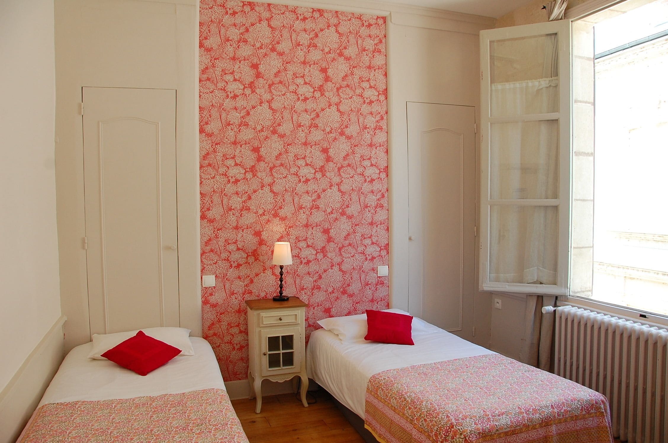 Una habitación con dos camas, una mesita de noche con lámpara, una ventana con cortinas y un radiador.