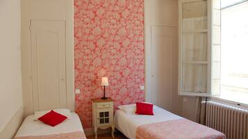 Una habitación con dos camas, una mesita de noche con lámpara, una ventana con cortinas y un radiador.