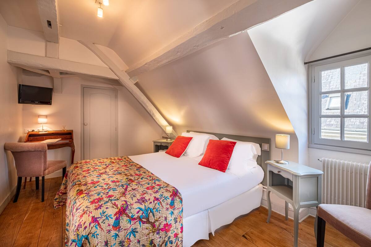 A bedroom with a bed, a desk, a chair, a TV, and a window with a view of a building.