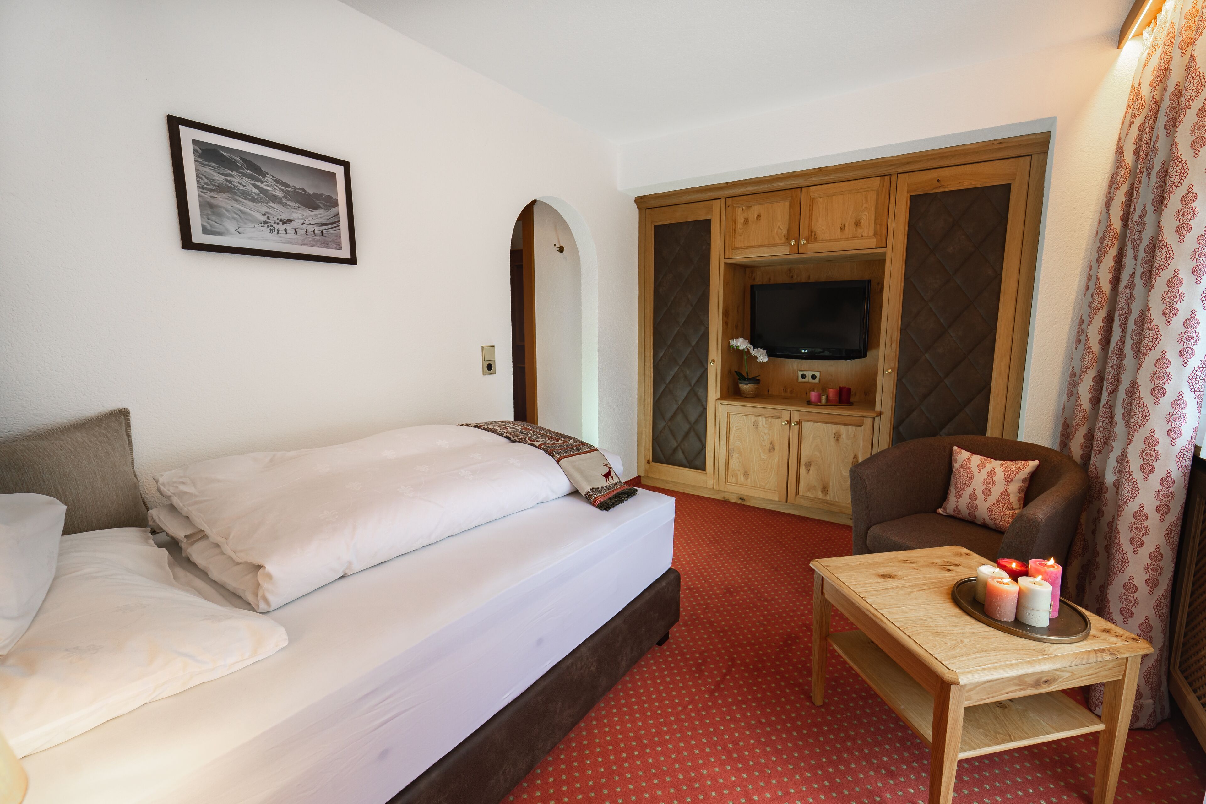 A bedroom with a bed, a wooden TV cabinet, a TV, a small table with candles, and a framed picture on the wall.
