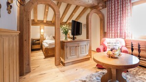 A wooden-floored room with a bed, a TV, a dining table with fruit, and a red chair. - Hotel Ulli (Lech am Arlberg)