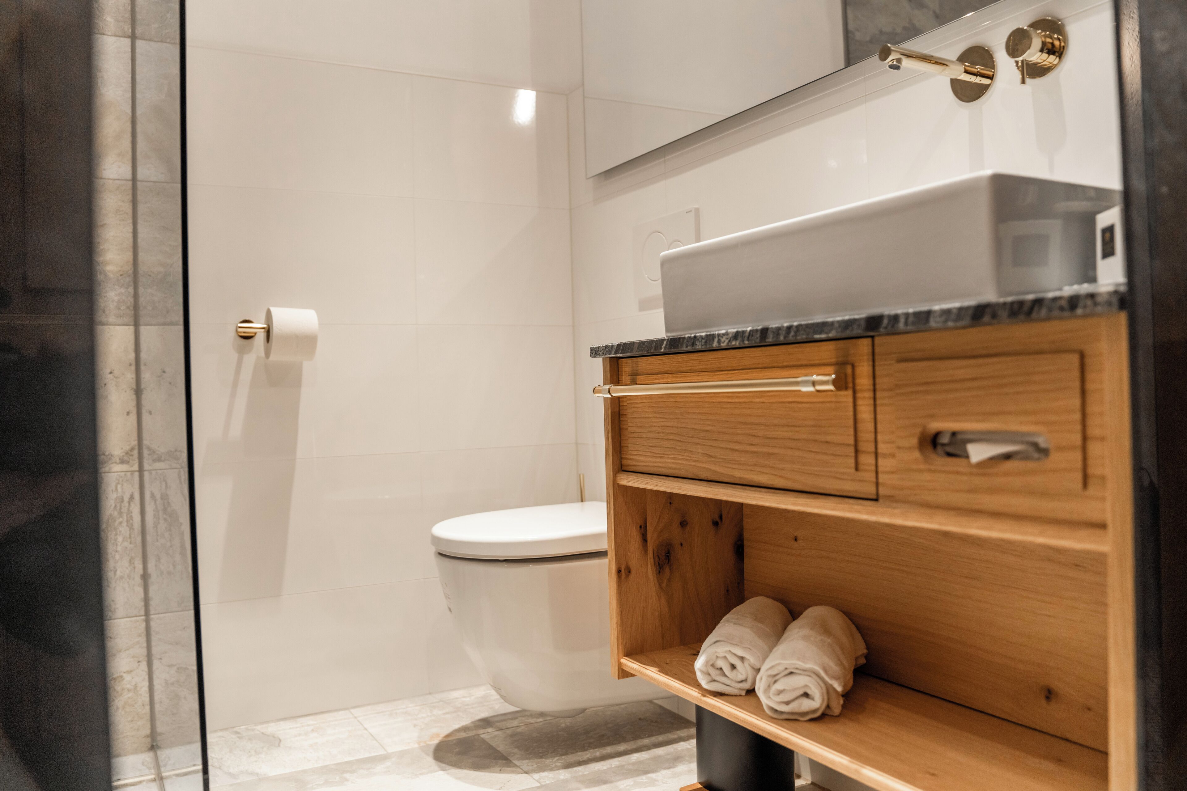 A modern bathroom with a glass shower enclosure, a white toilet, and a wooden vanity with a sink and towel storage.
