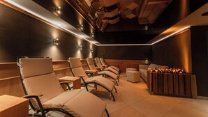 A modern spa room with multiple reclining chairs, a wooden side table, and a built-in fireplace. - Hotel Ulli (Lech am Arlberg)