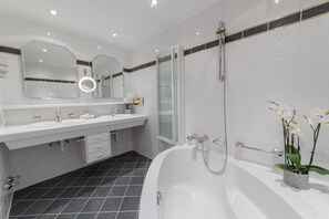 Un baño moderno con una bañera grande, dos lavamanos, cabina de ducha de vidrio y un espejo con luz circular.