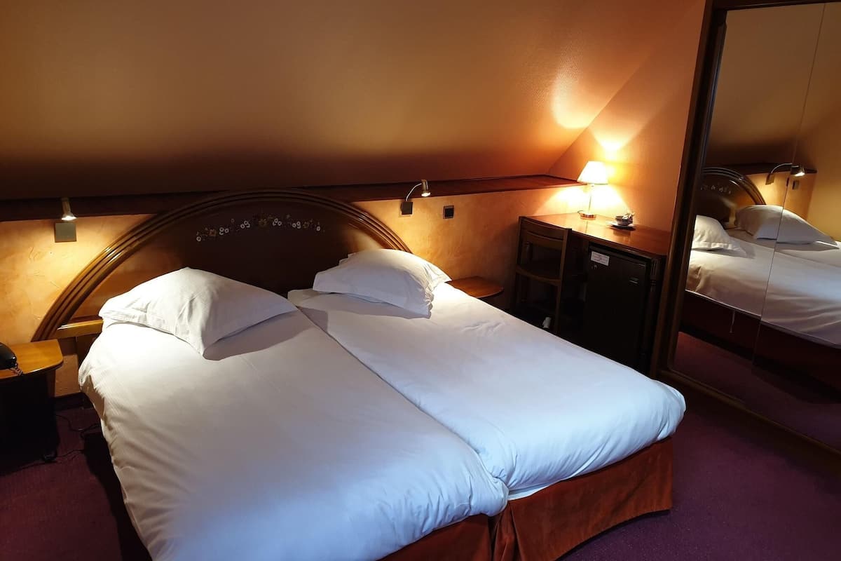 Een netjes opgemaakt bed met witte lakens, een houten hoofdbord, een nachtkastje met een lamp en een spiegel die het bed weerspiegelt.