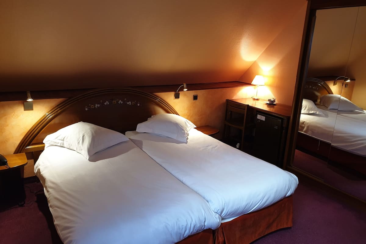 Een net opgemaakt bed met witte lakens, een houten hoofdbord, een nachtkastje met een lamp en een spiegel aan de muur.