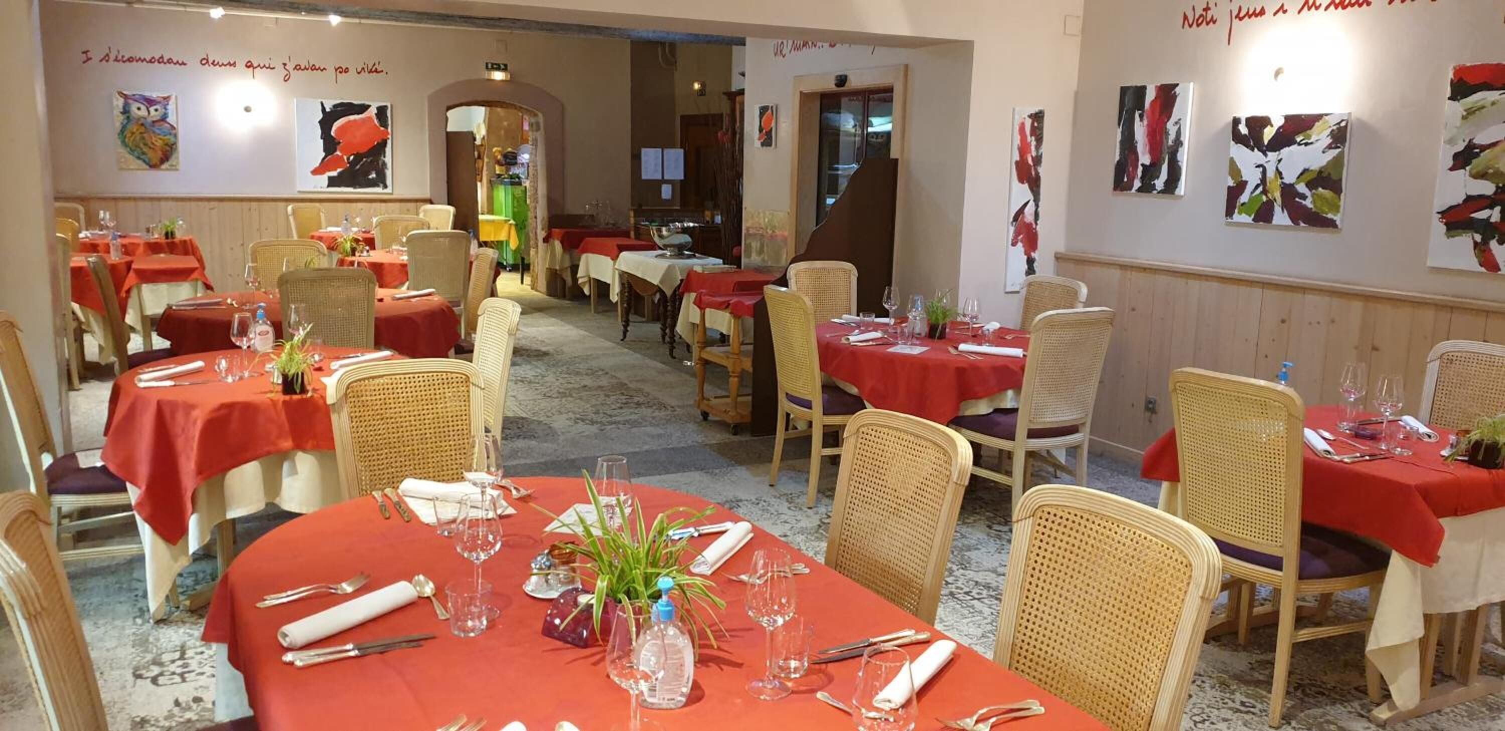 Un restaurante con varias mesas dispuestas para comer, con sillas de mimbre y manteles rojos.