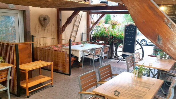 Ein Straßencafé mit Holztischen und -stühlen, einer Kreidetafel mit Speisekarte und Blumentöpfen.