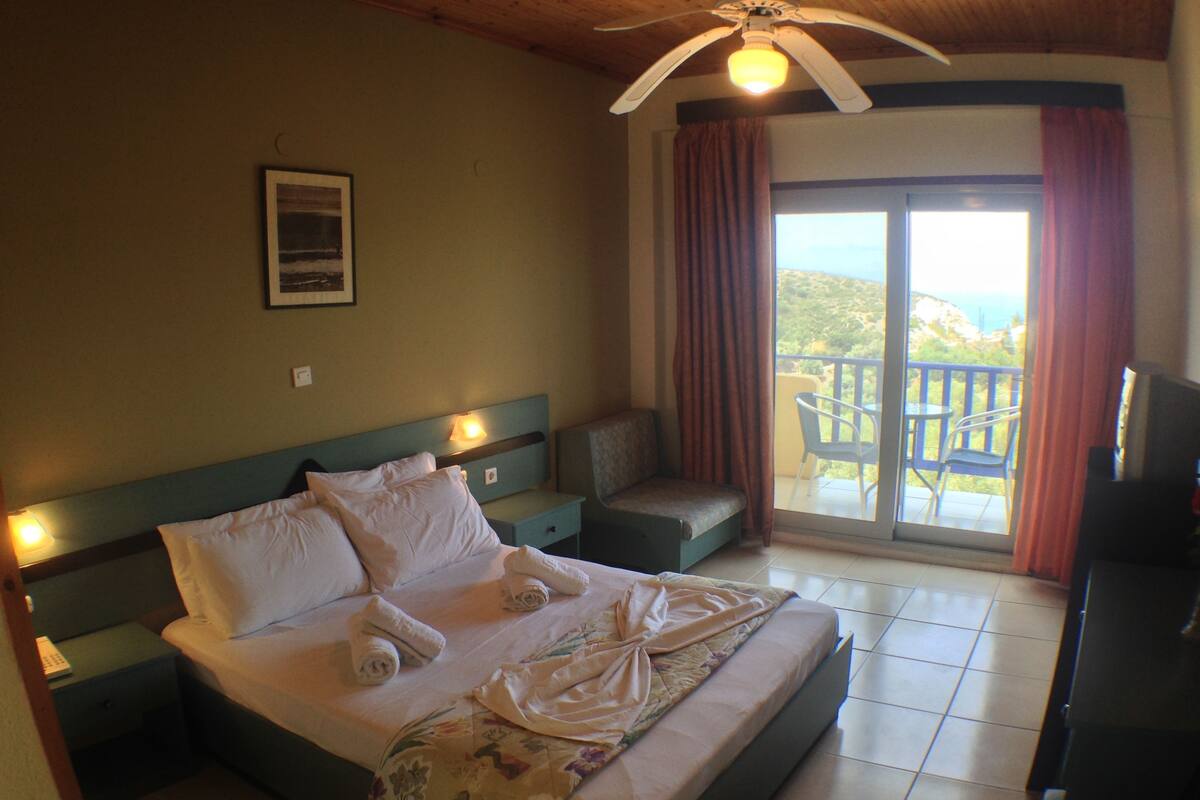 A hotel room with a bed, a nightstand, a chair, a TV, and a balcony with a view.