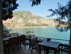 Une terrasse meublée de mobilier en bois, offrant une vue sur un lac, des bateaux et un paysage montagneux en arrière-plan.