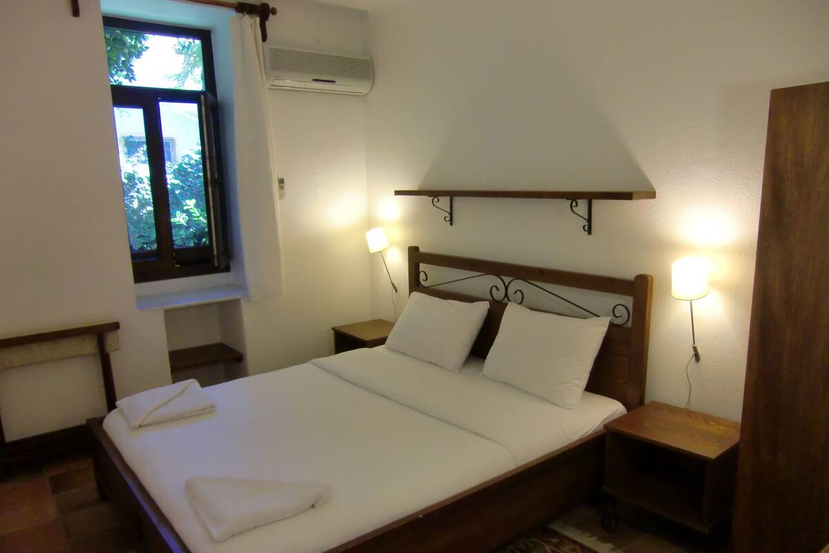A neatly made bed with white linens and a folded towel, two bedside tables with lamps, a window with curtains, and an air conditioning unit on the wall.