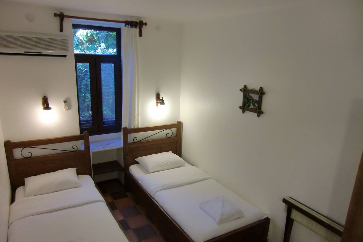 A room with two single beds, a window with curtains, a wall-mounted heater, and a small picture frame.