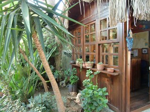 Une structure en bois surmontée d’un toit de chaume, entourée de différentes plantes en pot et de verdure.