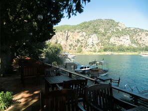 Une terrasse en bois, agrémentée de chaises et de tables, qui offre une vue sur un lac paisible, avec des bateaux amarrés à proximité et un paysage montagneux en arrière-plan.