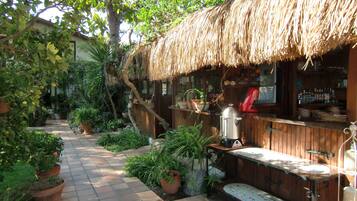 A rustic outdoor seating area with a thatched roof, surrounded by potted plants and a brick pathway.