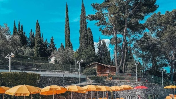 Piscina con tumbonas y sombrillas, rodeada de árboles y un muro de piedra.