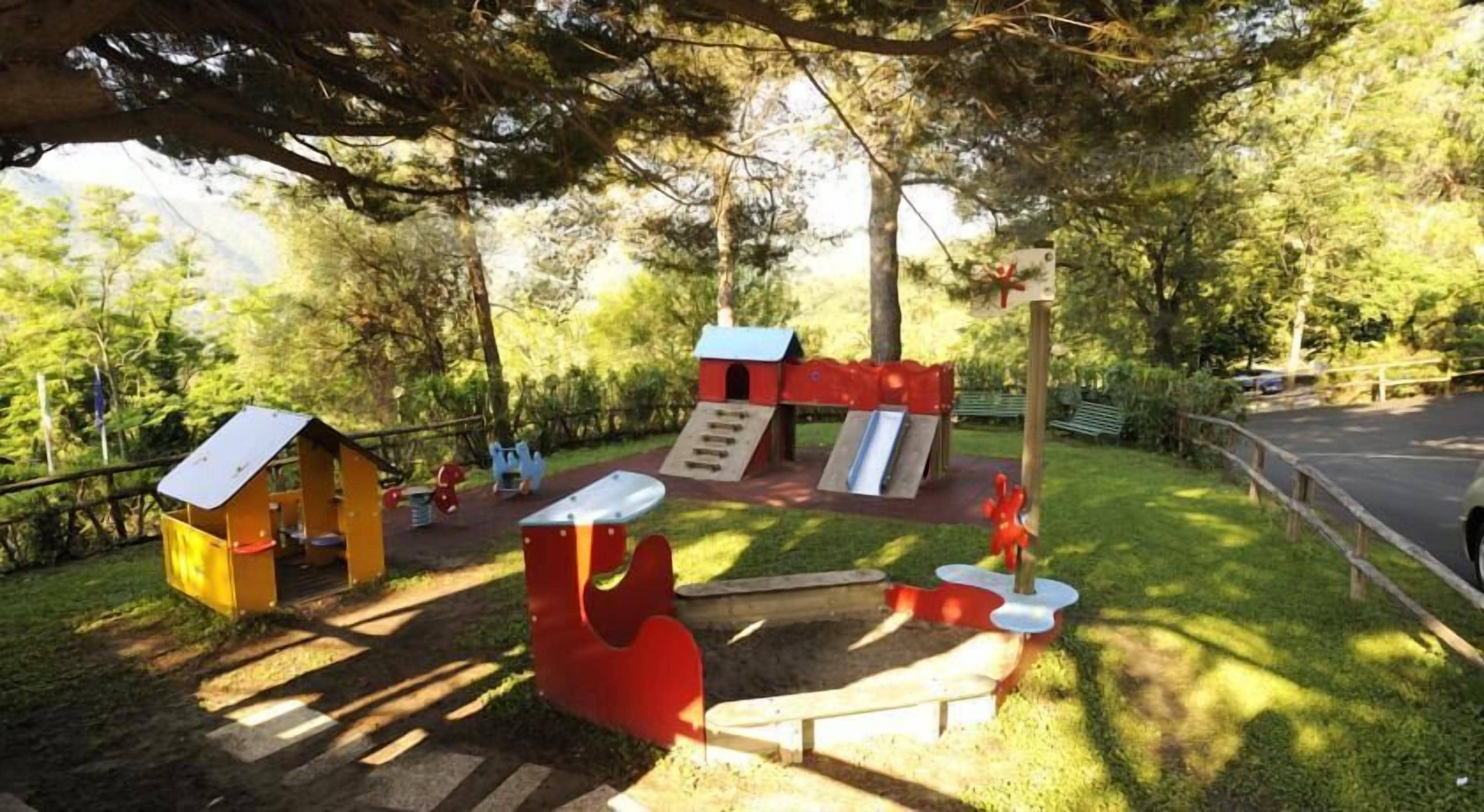 A playground with various play structures including slides and climbing areas, surrounded by trees and a paved path.