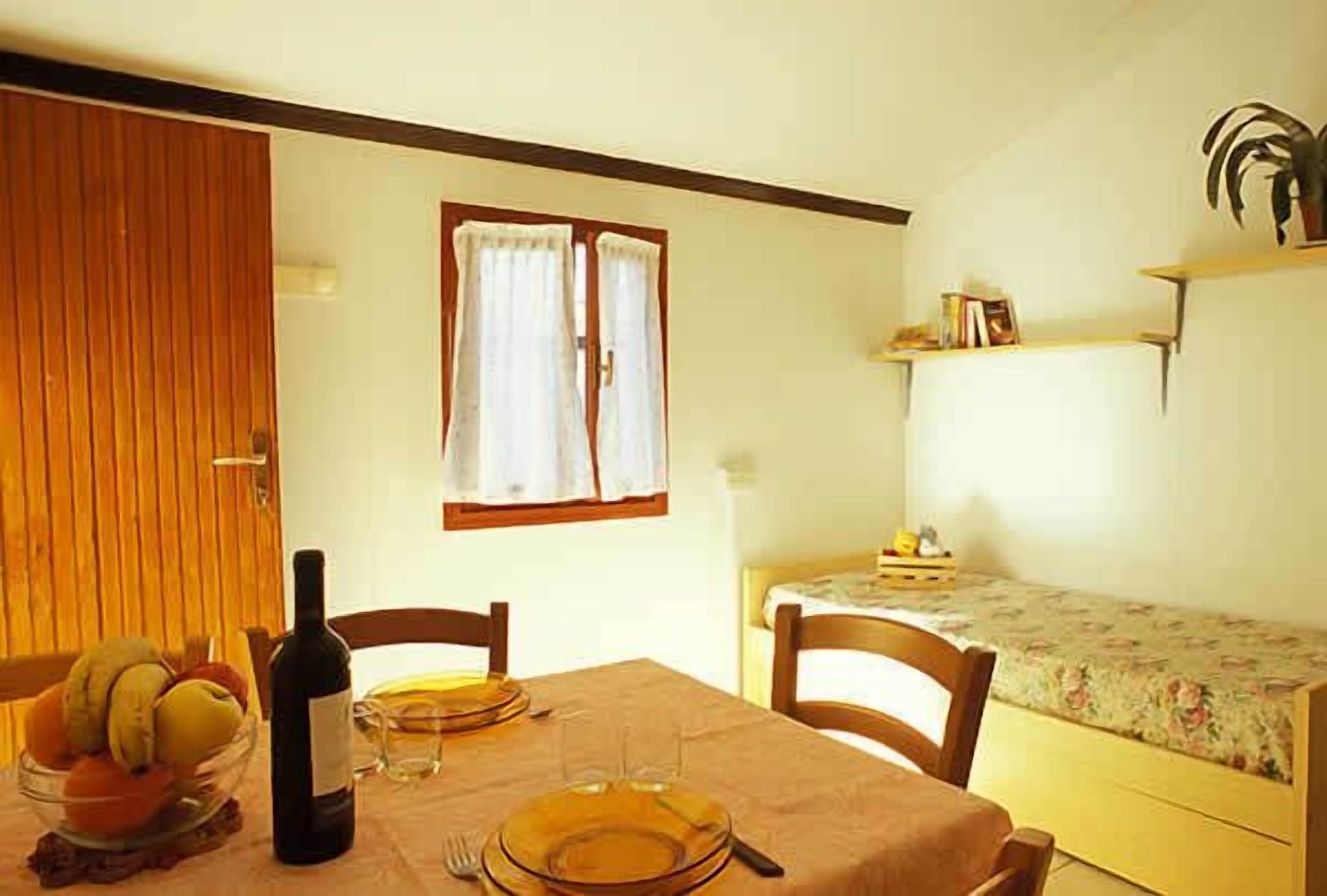 A dining area with a table set for two, a bottle of wine, and a bowl of fruit. There is a window with curtains, a bed with a floral bedspread, and a shelf with books and a plant.