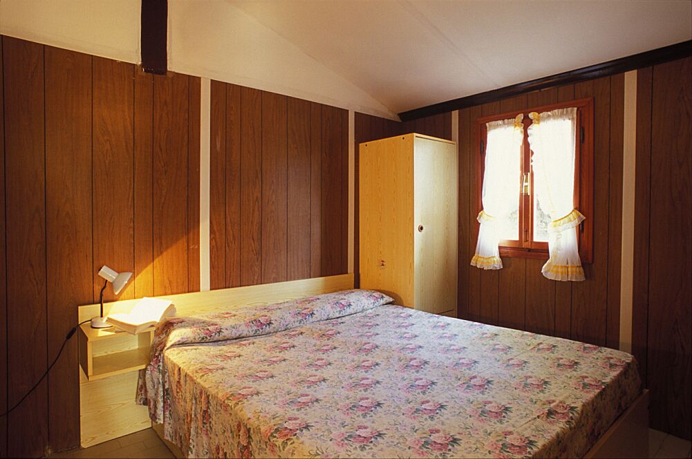 A bedroom with a wooden wardrobe, a bed with floral bedding, a nightstand with a lamp, and a window with curtains.