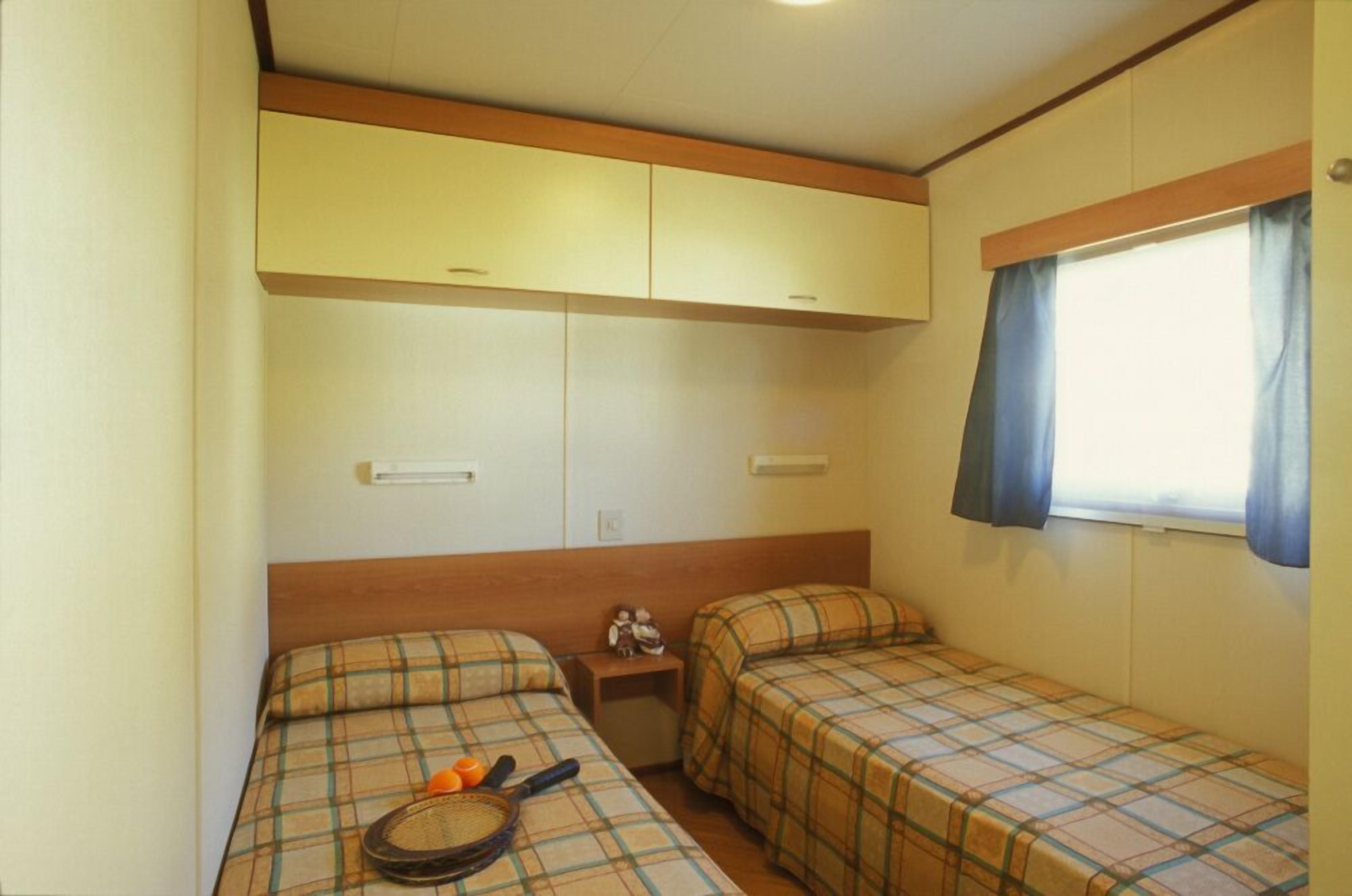 A small, compact room with two beds, a window with curtains, and a wooden headboard.