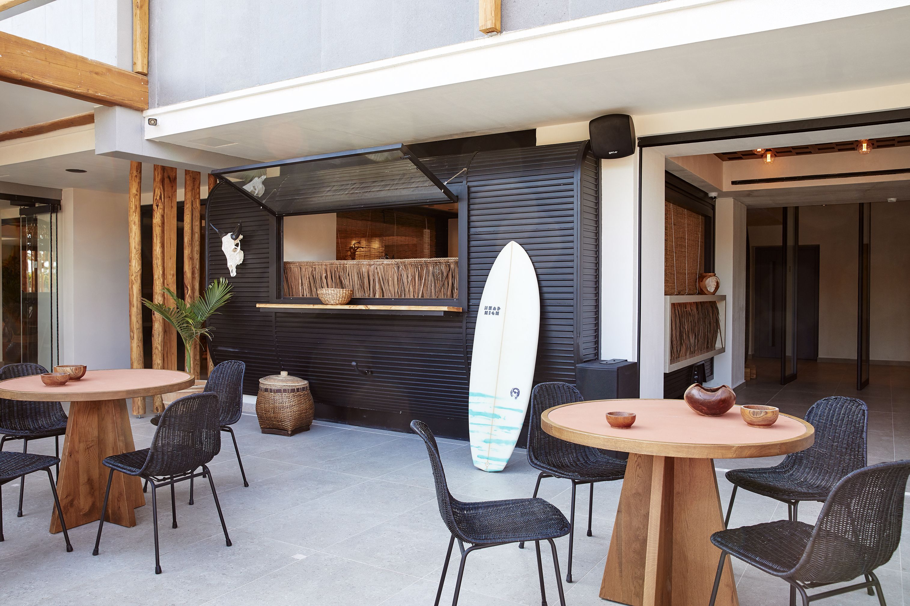 An indoor dining area with round wooden tables and black wicker chairs, a surfboard leaning against a wall, and a modern interior design.