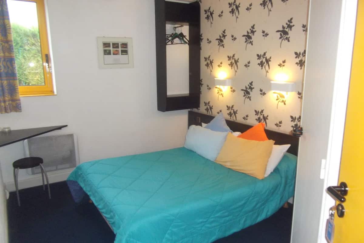 A small, neatly arranged bedroom with a bed, a desk, and a chair. There is a mirror on the wall, and the room has floral wallpaper.