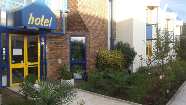 A hotel with a blue and yellow facade, a brick exterior, and a small garden area with various plants and flowers. - Rev'Hotel Coignières (Coignieres)