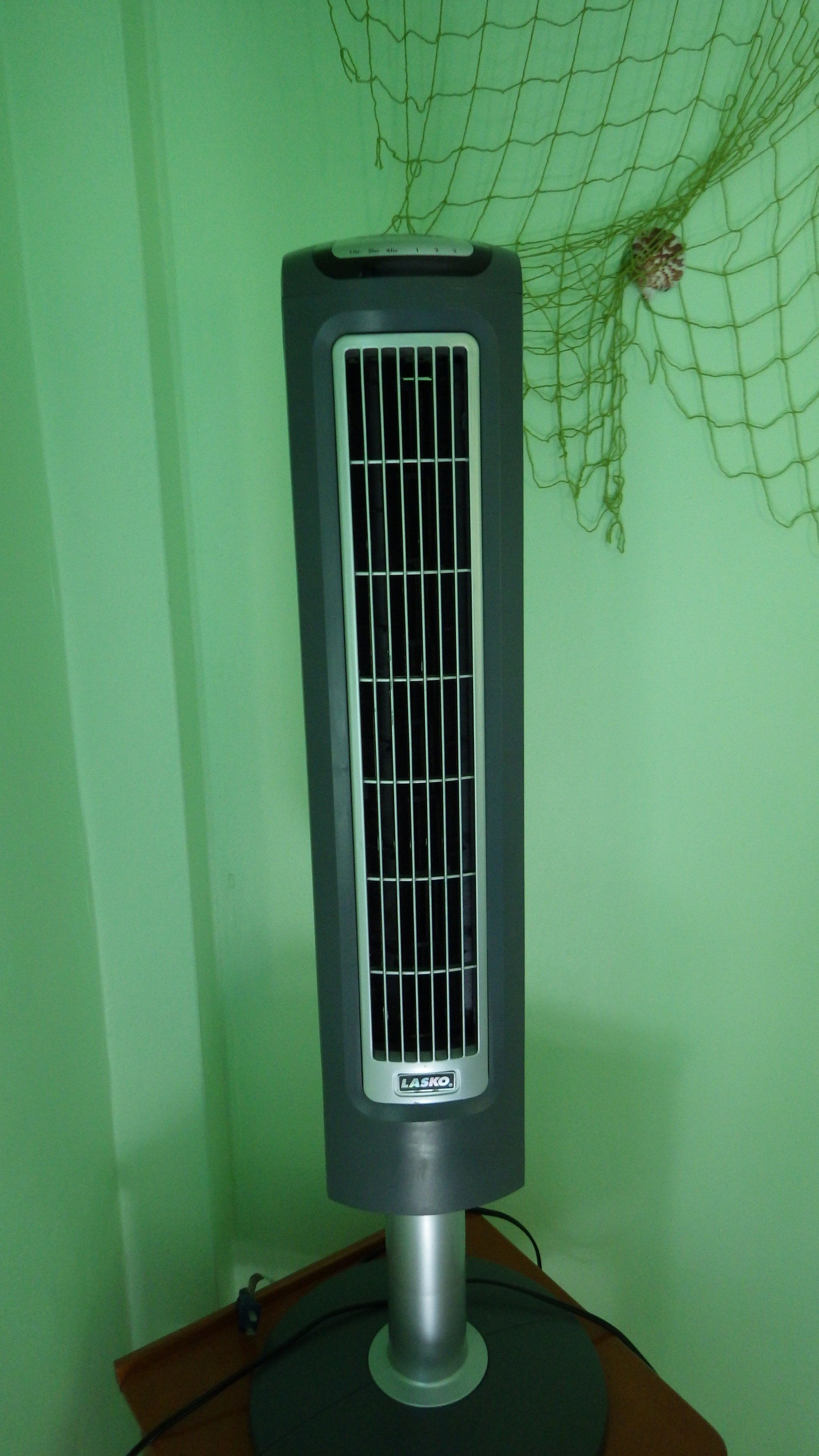 A standing fan with a control panel, placed on a table against a green wall.