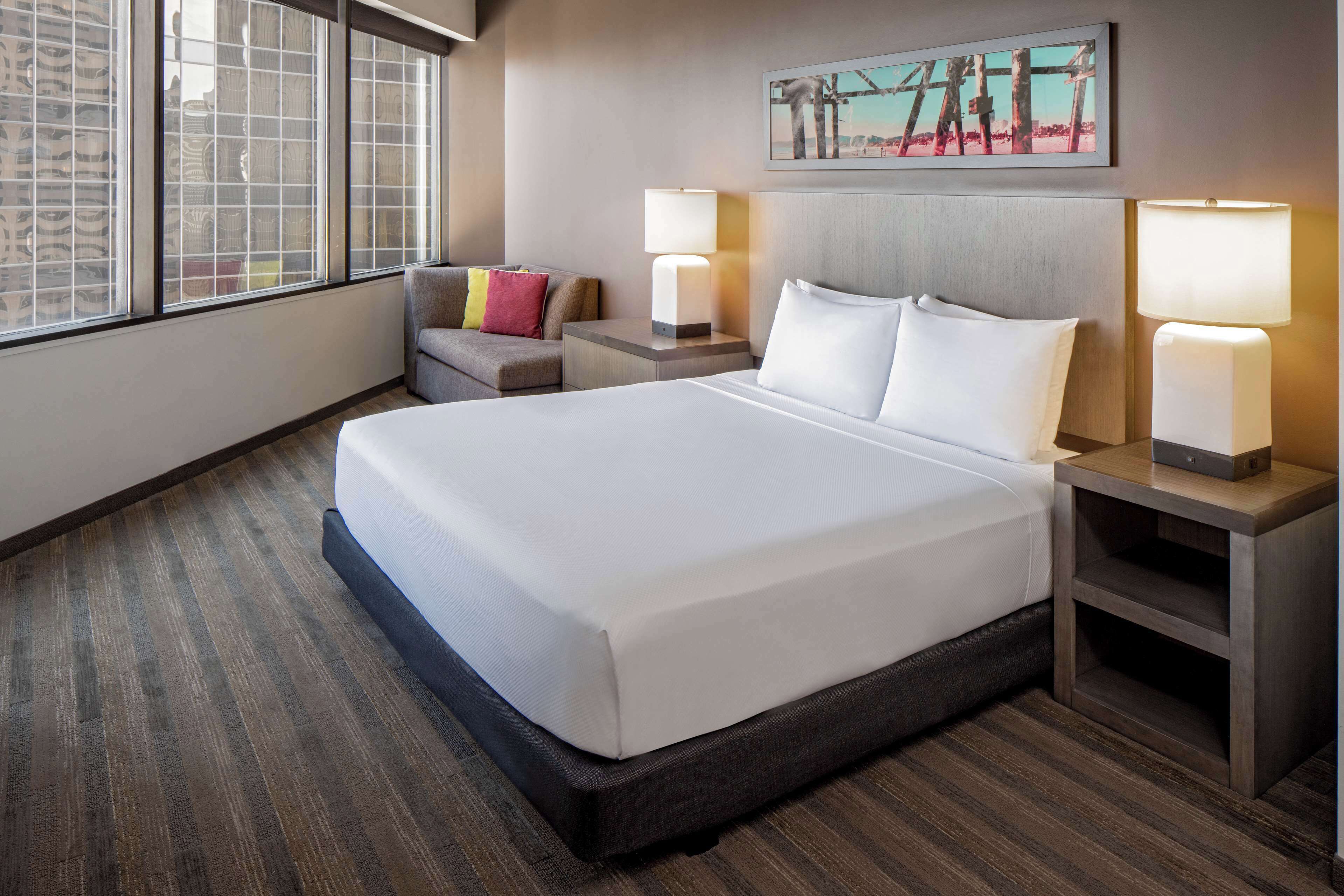 A hotel room with a large bed, two bedside tables with lamps, a small sofa with cushions, and a large window with a view.