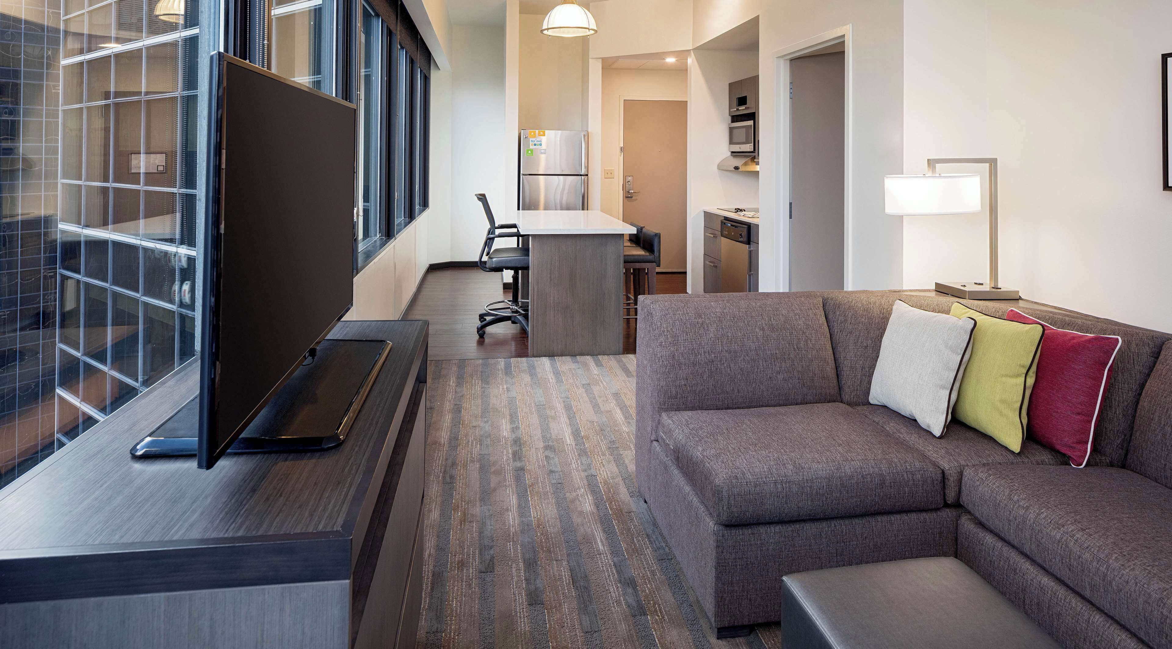 Una habitación de hotel moderna con un ventanal grande, un sofá con cojines de colores, un escritorio con una computadora portátil y una zona de cocina al fondo.