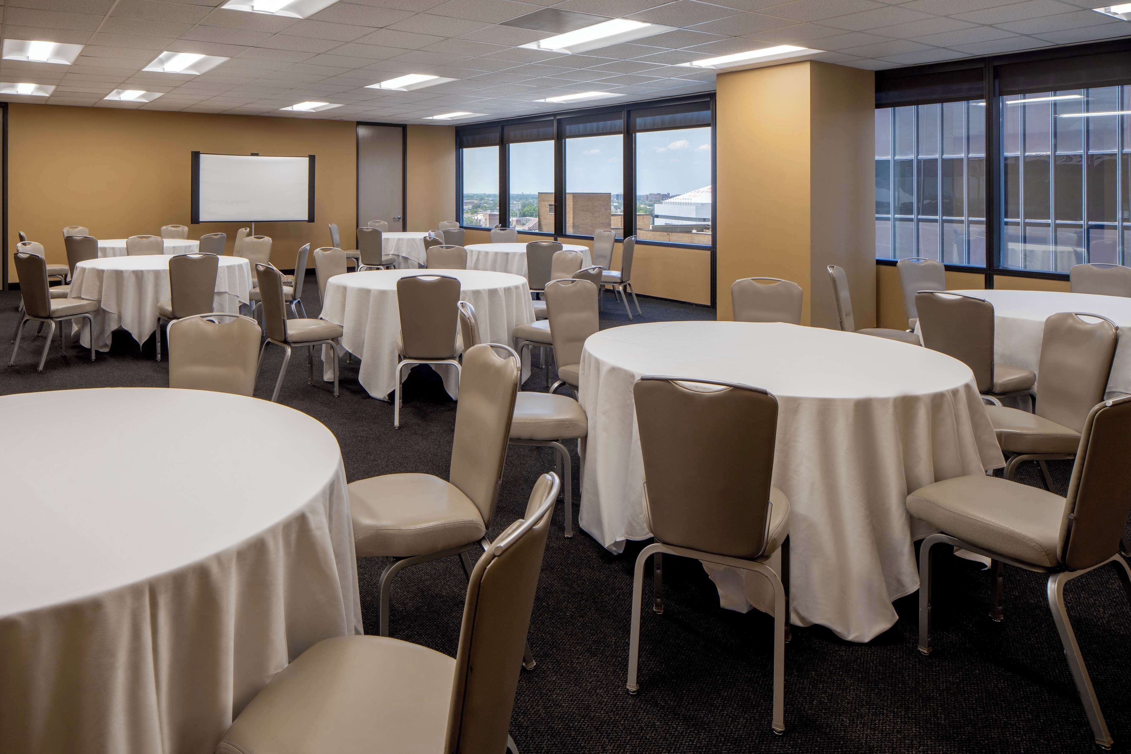 A conference room with multiple round tables and chairs, large windows with a view, and a projection screen on the wall.