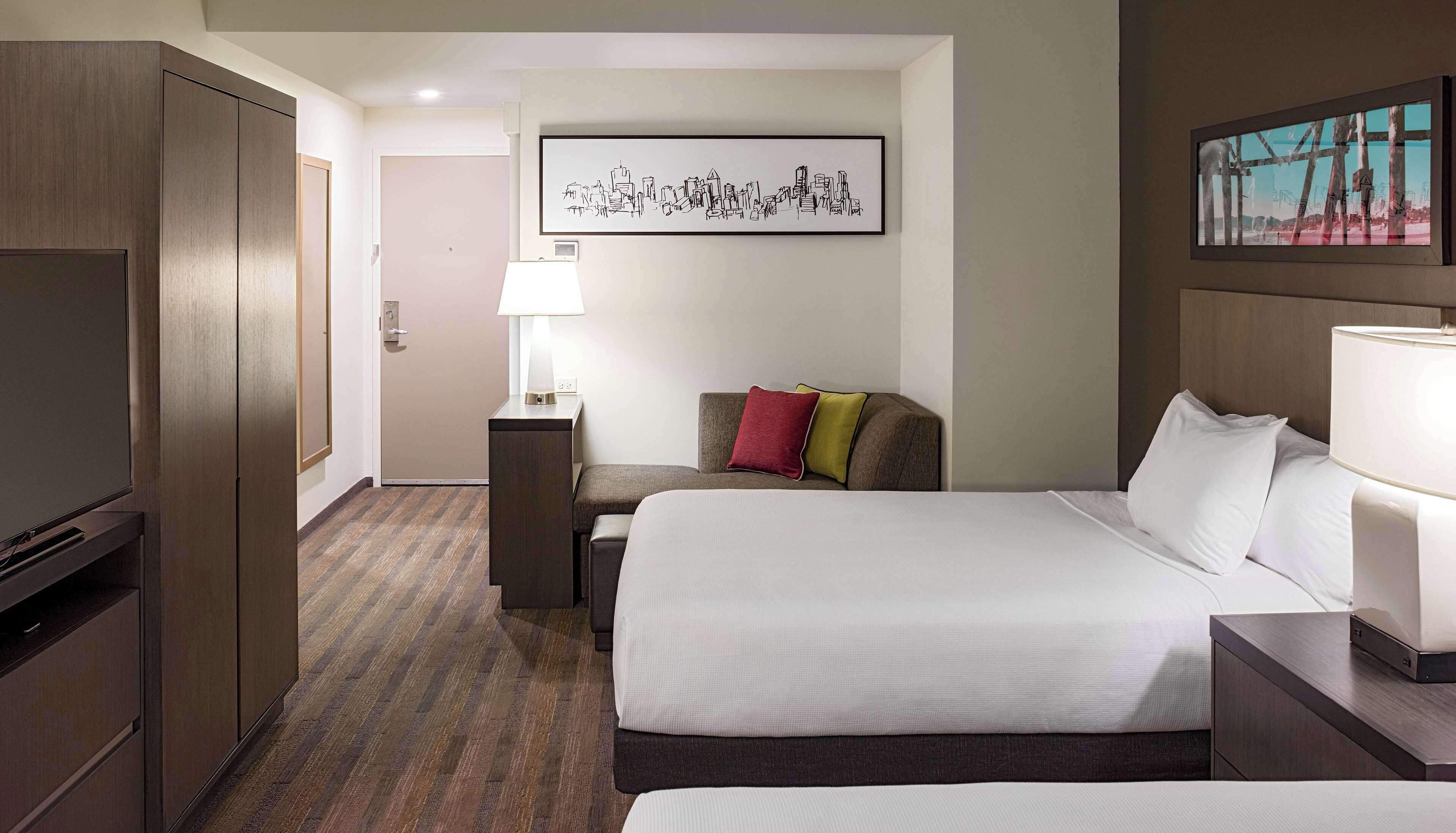 A hotel room with a large bed, a desk, a chair, a TV, and a framed cityscape picture on the wall.