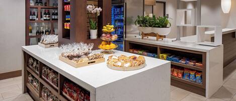A reception area with a counter displaying pastries, snacks, and bottled beverages. There are shelves with packaged goods and a potted plant on the counter.