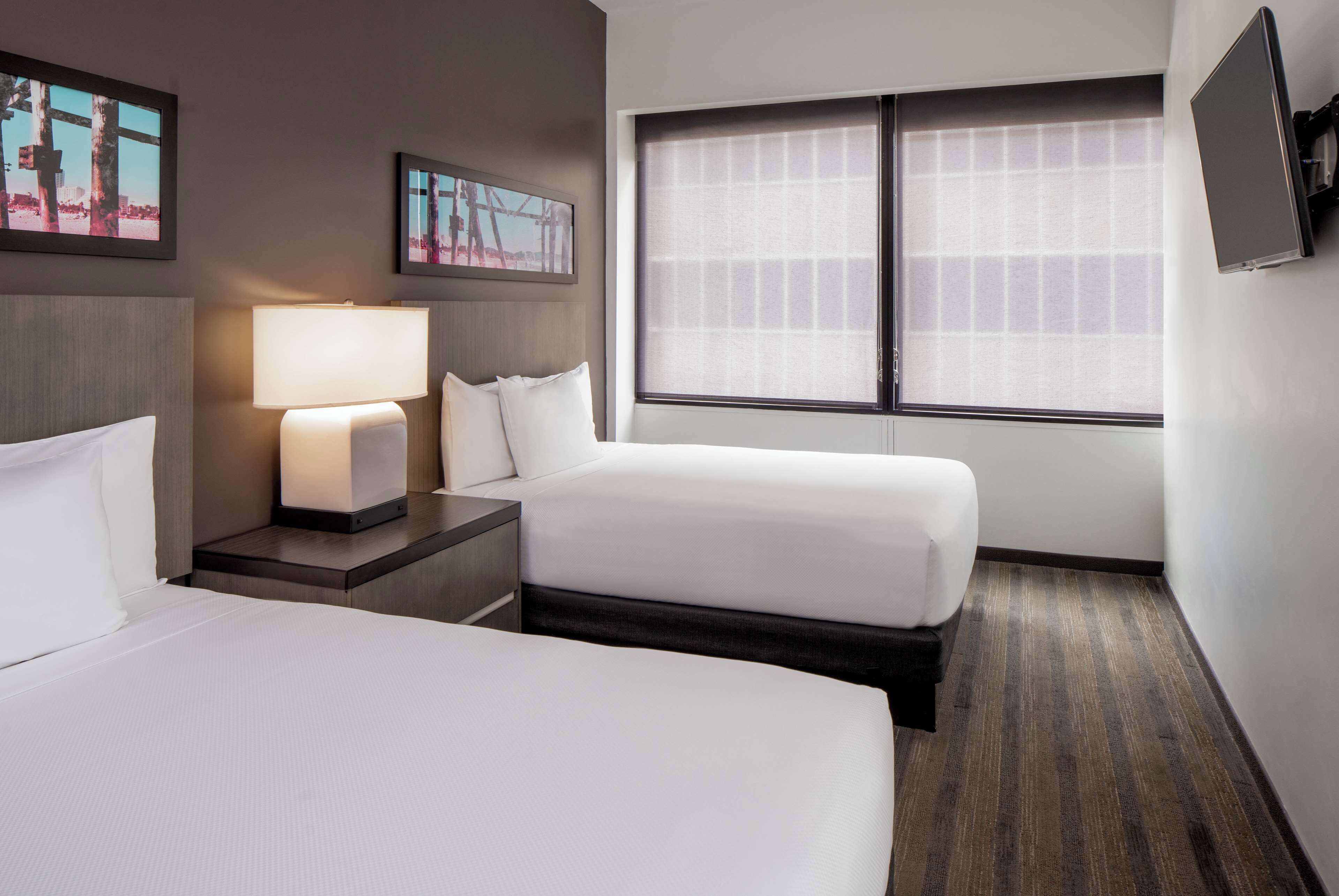 A hotel room with two beds, a nightstand with a lamp, a large window with blinds, and framed pictures on the wall.