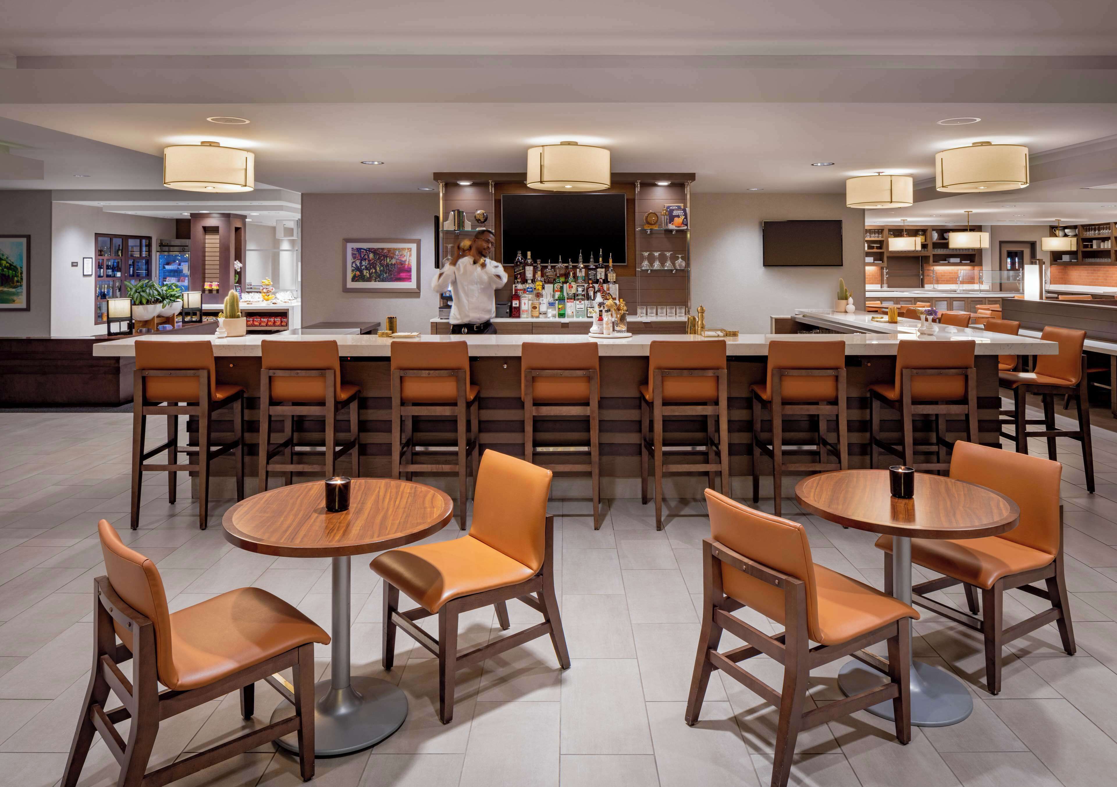 A modern bar area with multiple seating options including round tables and bar stools, a well-stocked bar counter, and ambient lighting.