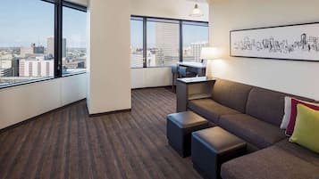 A modern hotel room with a large window offering a city view, a comfortable sofa, a desk with a chair, and a cityscape artwork on the wall.