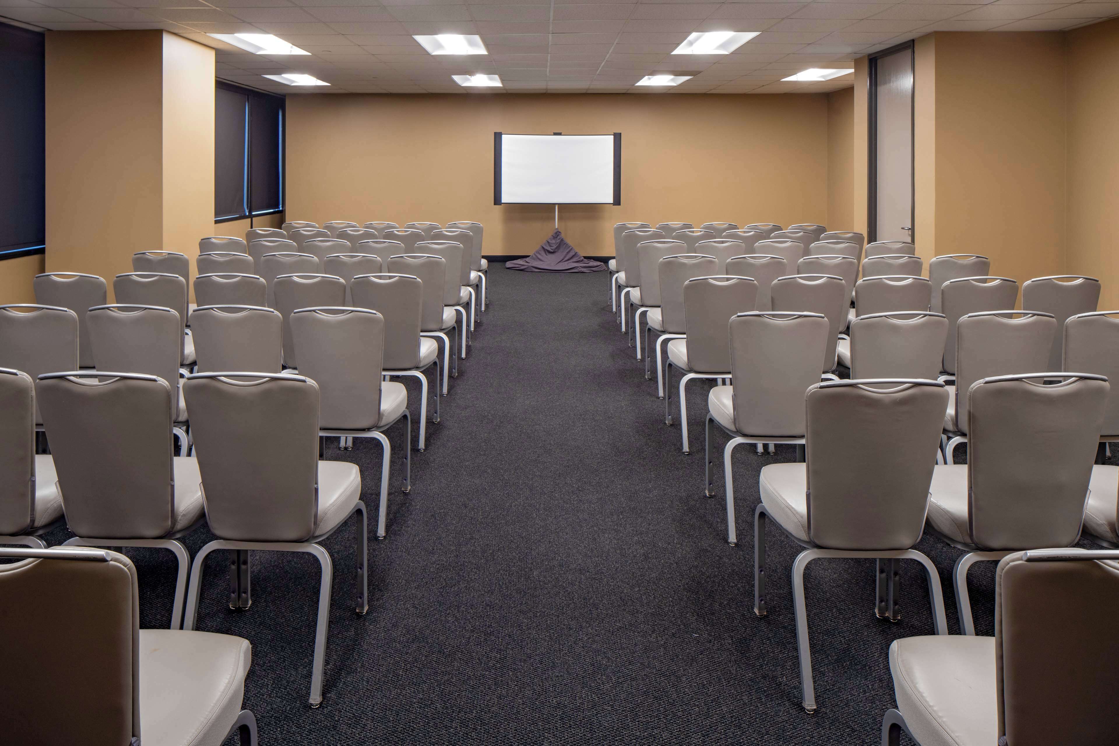 Uma sala de conferência com fileiras de cadeiras voltadas para uma tela de projeção vazia.