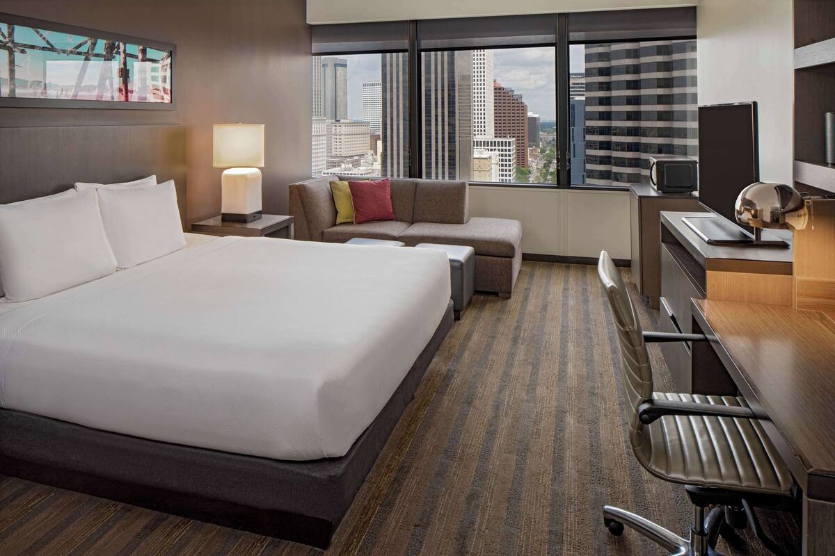 Une chambre d’hôtel avec un grand lit, un bureau avec un ordinateur, une chaise, un canapé et une fenêtre donnant sur la ville.