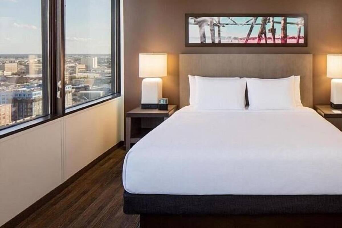 Une chambre d’hôtel avec un grand lit, deux tables de chevet, une commode, une fenêtre donnant sur la ville et un tableau au mur.