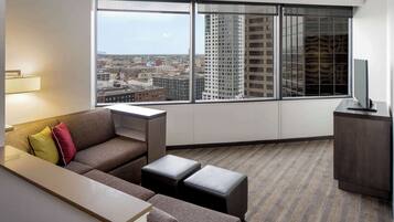 A modern hotel room with a large window, a sofa, two ottomans, a small table, and a TV on a cabinet.