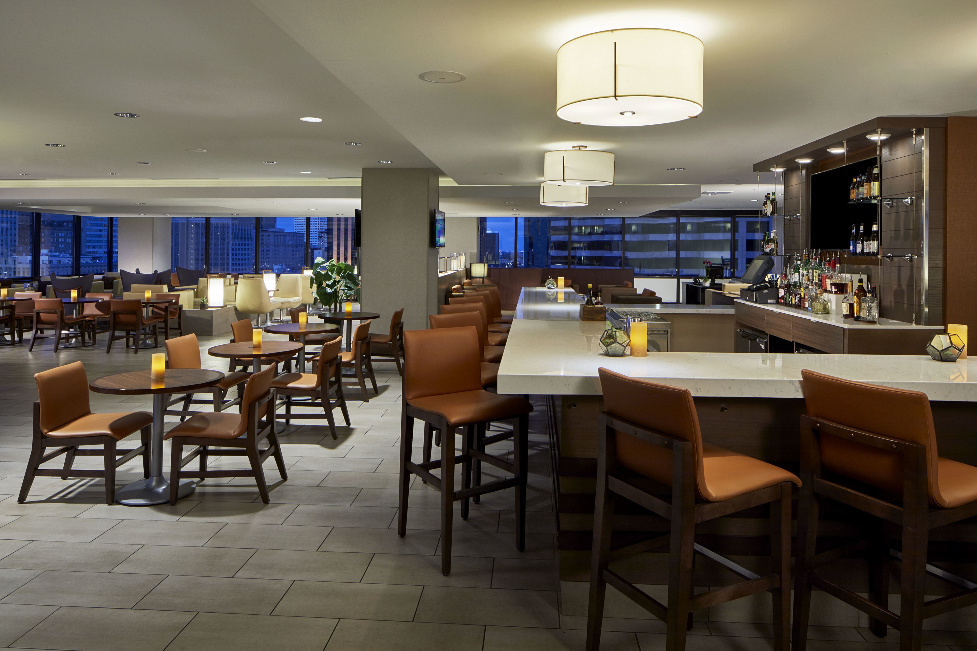 A modern hotel lobby with multiple seating areas, a bar, and a large window offering a city view.