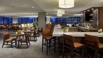A modern hotel lobby with multiple seating areas, a bar, and a large window offering a city view.