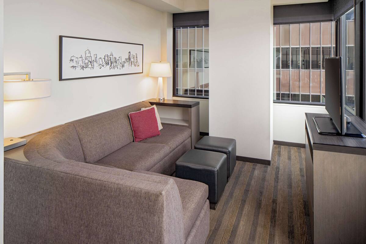 Une chambre d’hôtel moderne avec un canapé gris, un coussin rouge, une lampe sur pied, un téléviseur à écran plat et une œuvre d’art représentant un paysage urbain, encadrée et accrochée au mur.
