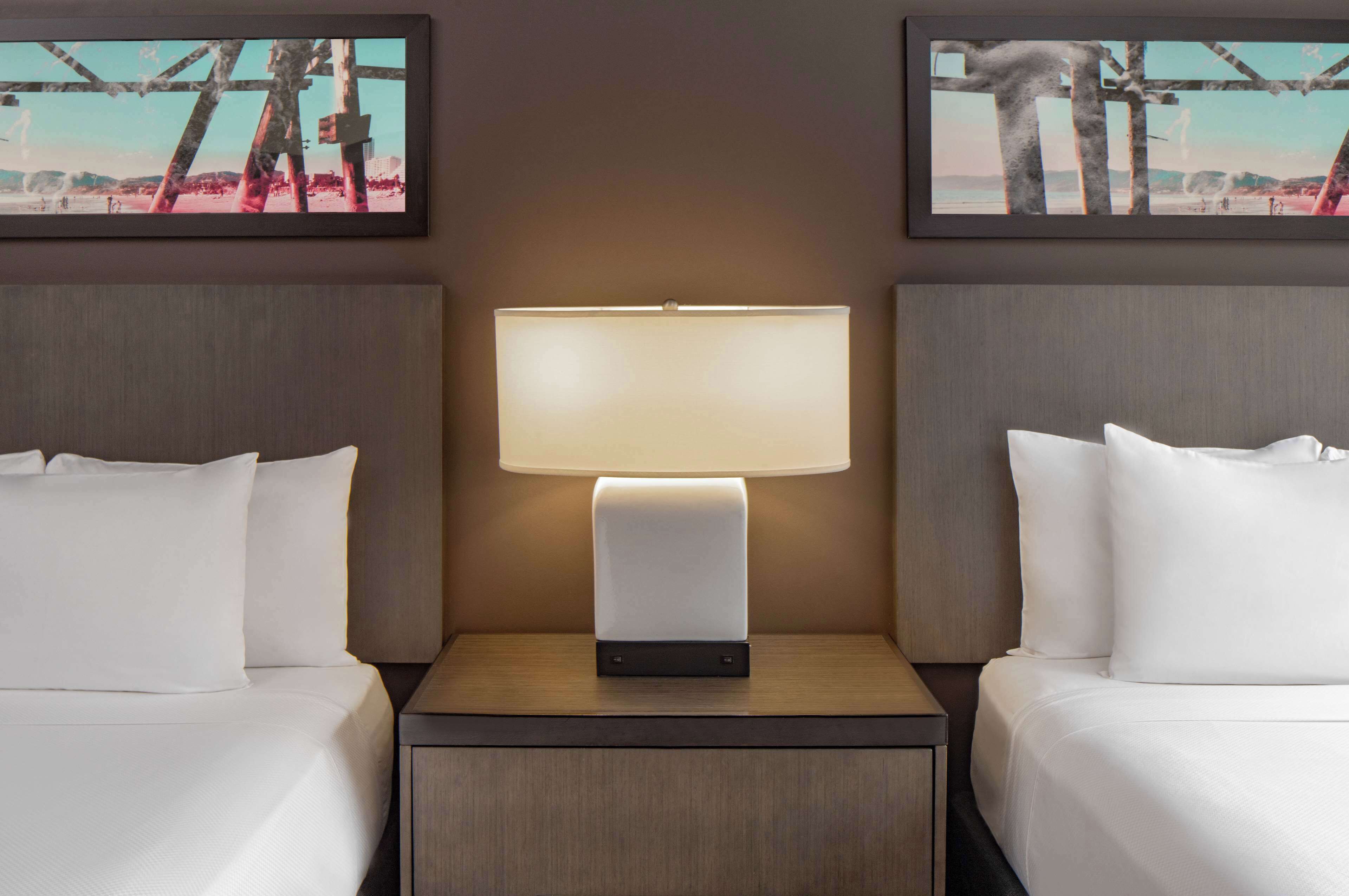 A hotel room with two beds, a nightstand with a lamp, and two framed pictures on the wall.