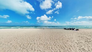 Une vaste plage de sable fin, sous un ciel bleu dégagé, avec des vagues qui déferlent sur l’océan. On y voit plusieurs chaises de plage et un petit véhicule garé sur le sable.