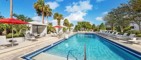 A rectangular swimming pool with clear blue water, surrounded by lounge chairs and umbrellas, with palm trees and a clear sky in the background.