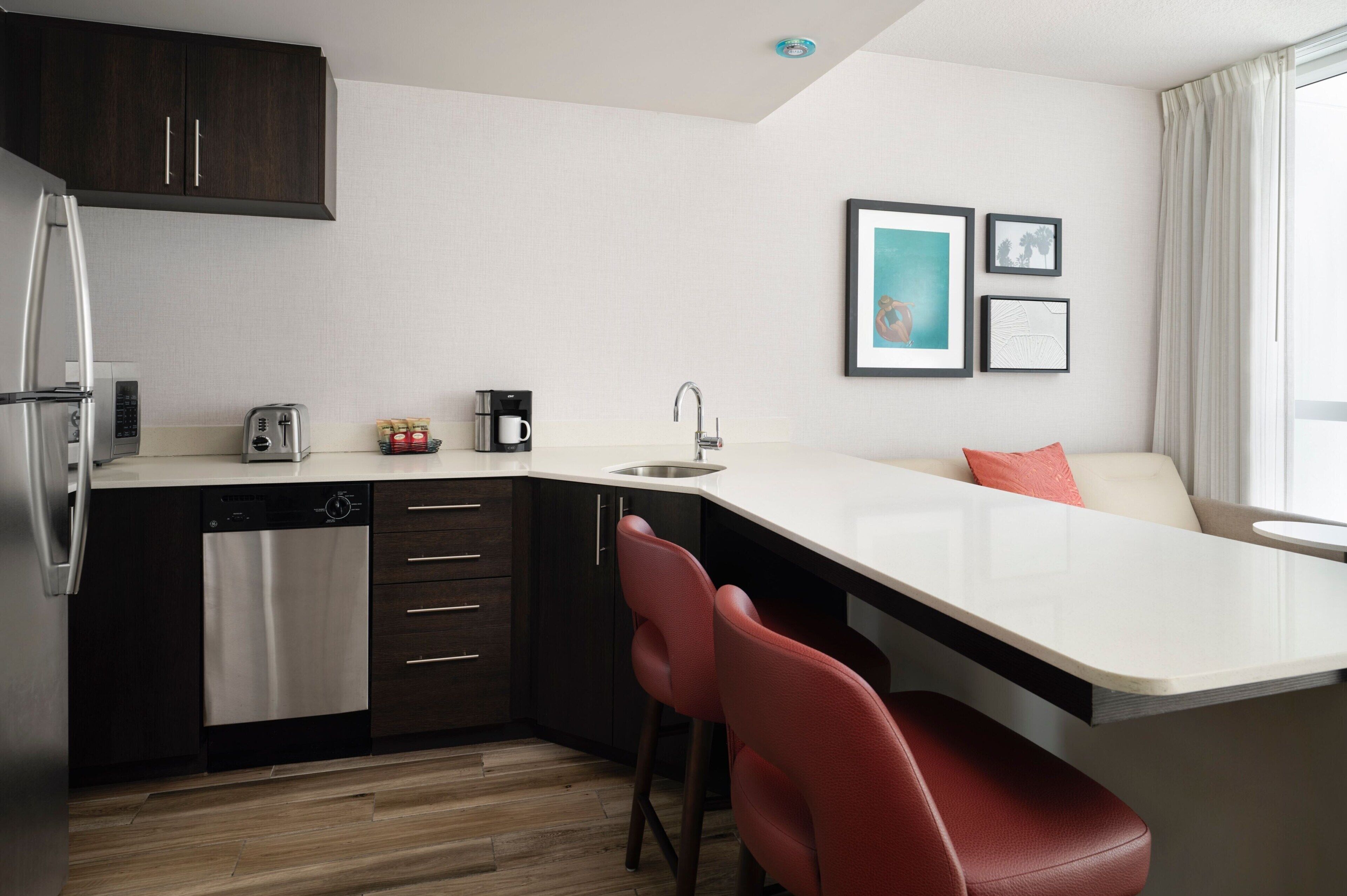 A modern kitchen with a dishwasher, a coffee maker, and a toaster. There is a sink with a faucet, a countertop, and a chair. The room has a window with curtains and two framed pictures on the wall.