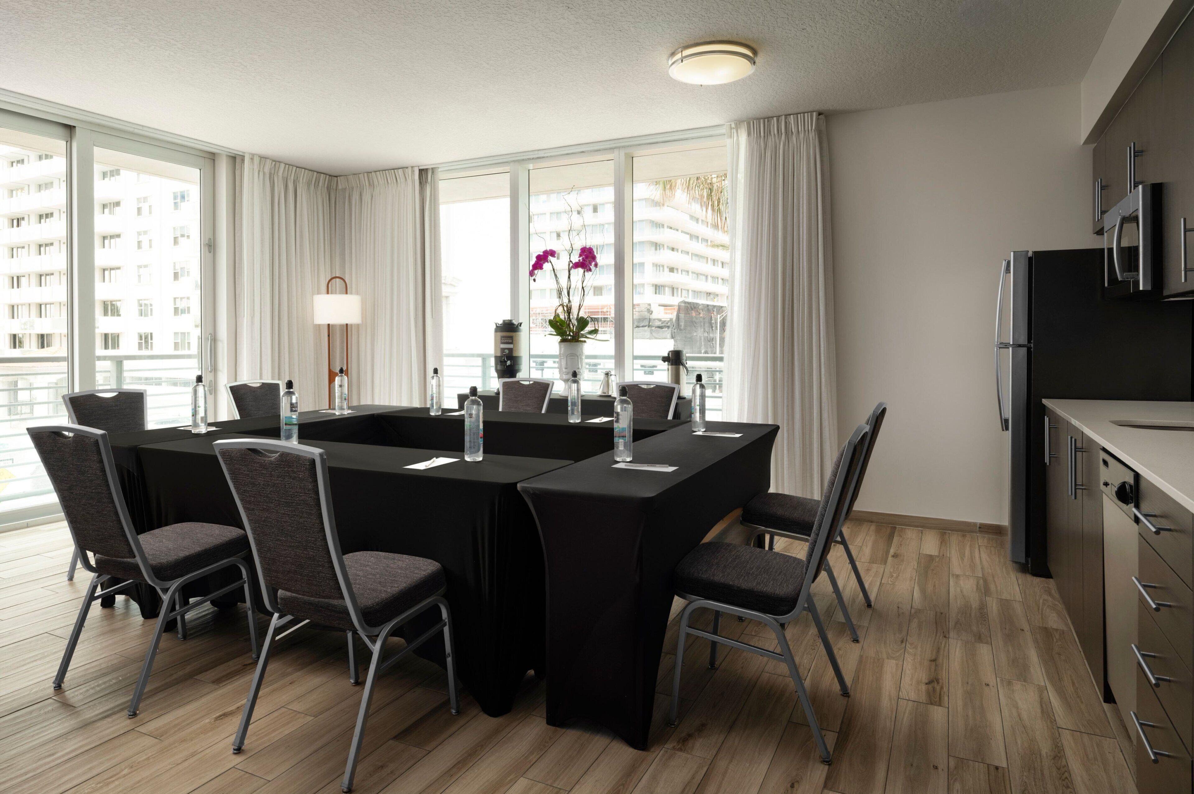 Une chambre d’hôtel moderne dotée d’une table à manger, de chaises, d’un coin cuisine et de grandes fenêtres offrant une vue sur la ville.