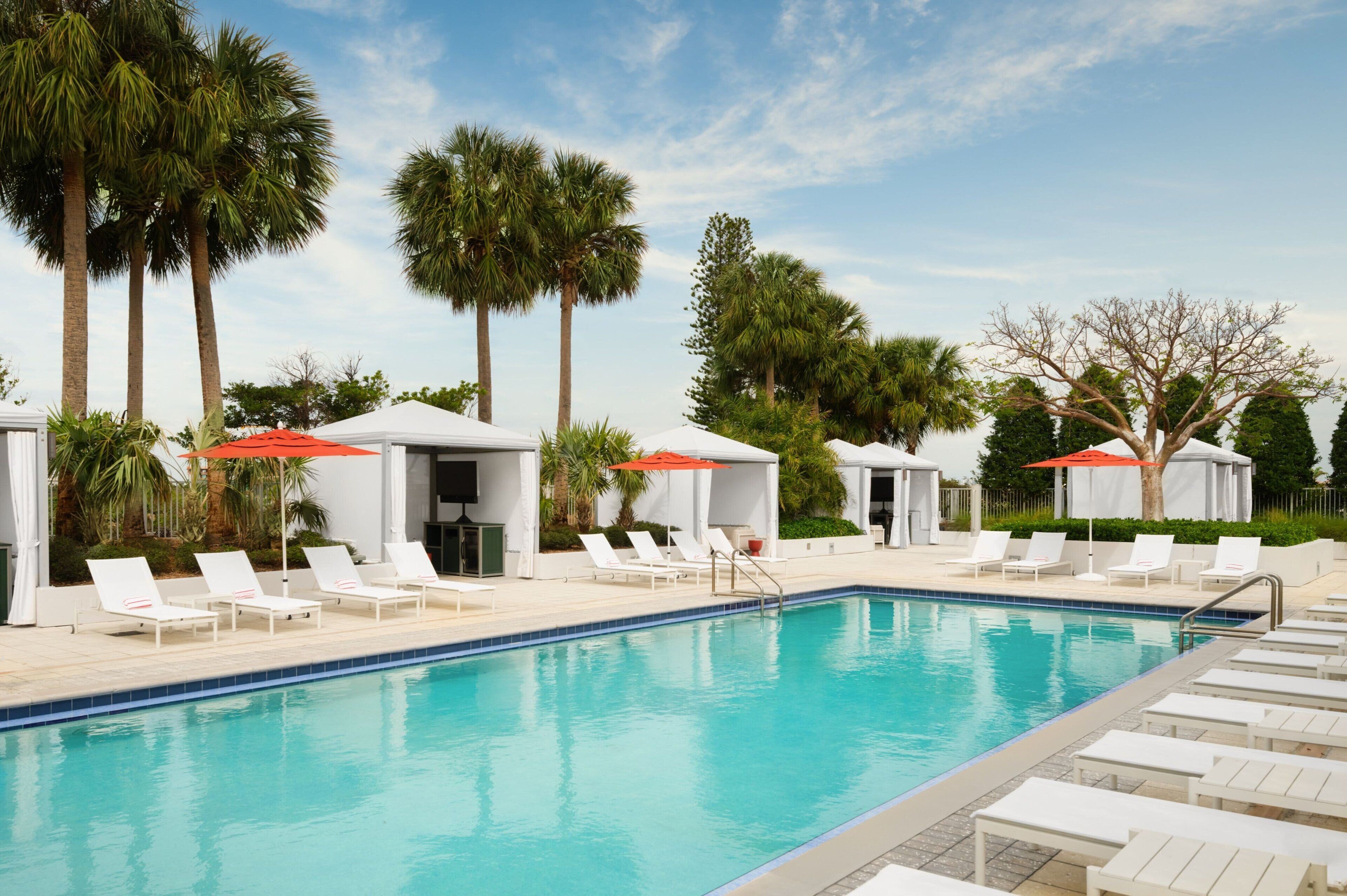 A swimming pool with white lounge chairs and red umbrellas, surrounded by palm trees and covered seating areas.