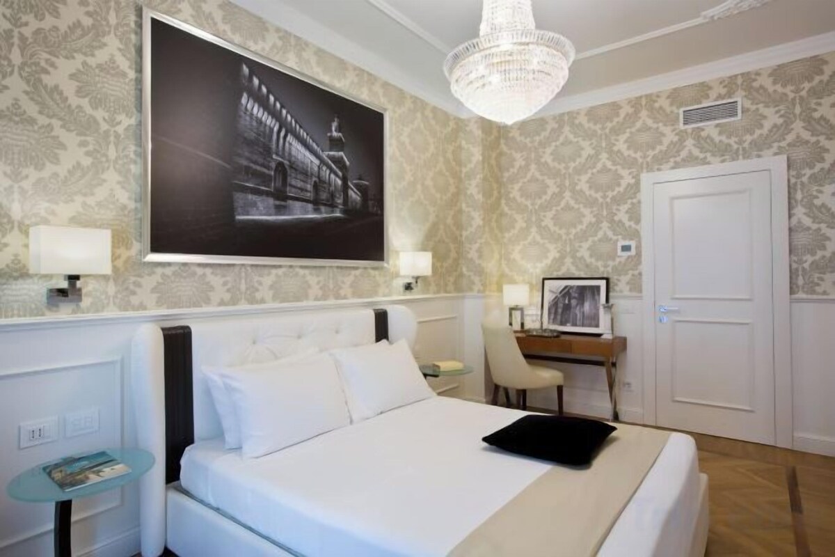 A hotel room with a large bed, a desk, a chair, a chandelier, and a framed picture on the wall.